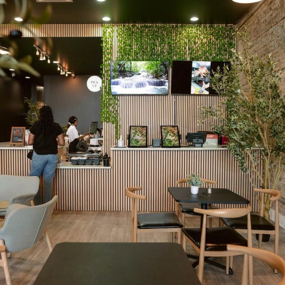 Modern restaurant interior with wood furnishings, exposed brick wall, greenery, counter service, and customers seated near tables.